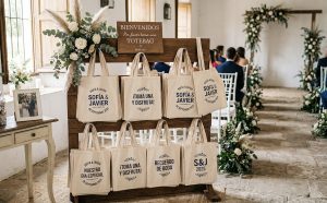 Un expositor de madera rústico en una ceremonia de boda, decorado con pampas y flores blancas, que sostiene filas de tote bags de tela personalizadas para los invitados. Las bolsas de lona de color crema tienen diseños impresos en azul con los nombres "SOFÍA & JAVIER" y frases como "¡TOMA UNA Y DISFRUTA!" y "NUESTRO DÍA ESPECIAL", listas como recuerdos de boda útiles.